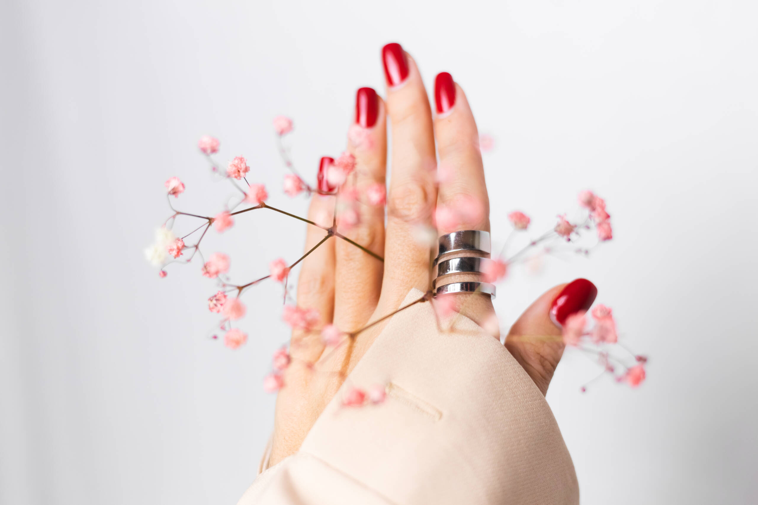 Mani femminili con manicure rossa ed elegante anello, fotografate con piccoli fiori rosa.