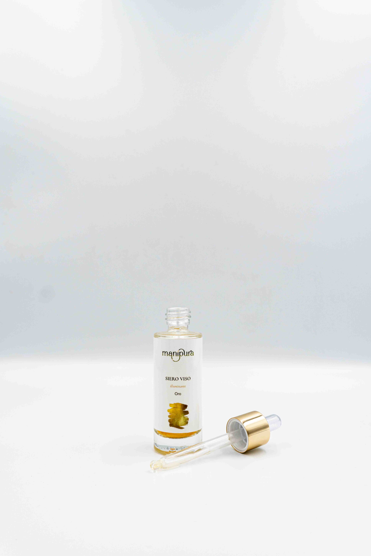 Boccetta aperta del siero viso illuminante Oro di Manipura con contagocce dorato appoggiato su sfondo bianco.